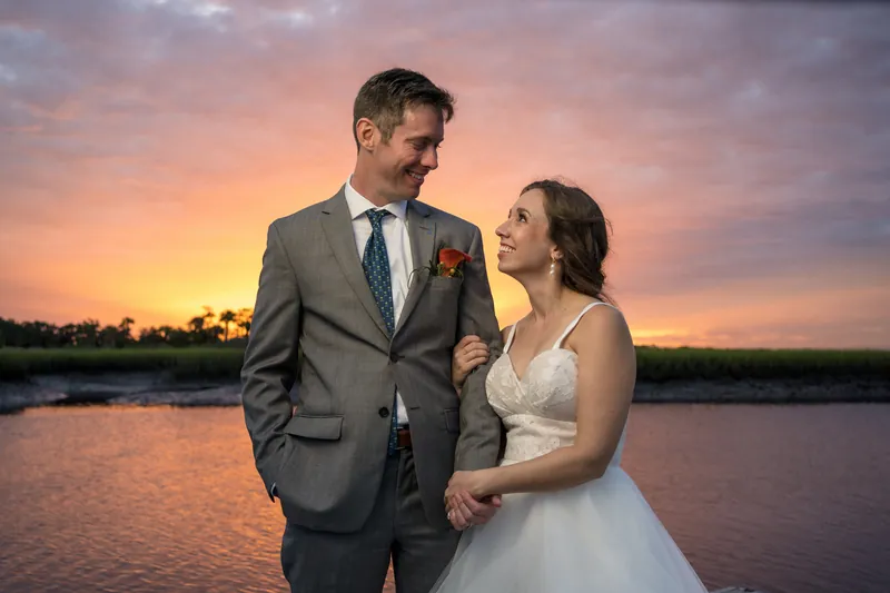 Kyle and Emma face each other on a dock at sunset, with golden light reflecting off the water behind them.