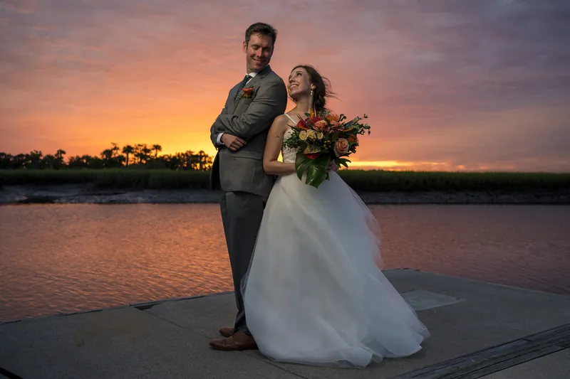 Kyle and Emma stand on a wooden deck overlooking the water at golden hour, Emma holding her bouquet as the sky burns orange