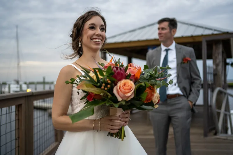 Emma holds a coral and red rose bouquet while Kyle stands nearby at Oyster Bay Yacht Club, a bridge visible behind them.