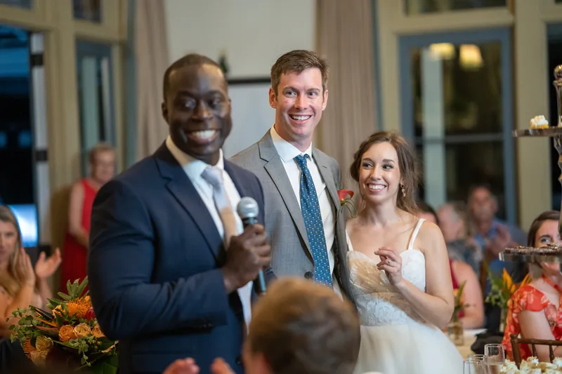 Two men and Emma laugh together during toasts at the reception, with guests seated at tables in the background.