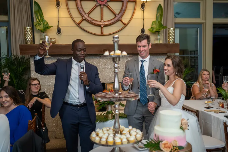 A man speaks at the microphone while Kyle and Emma stand beside him, surrounded by guests and white lantern centerpieces.