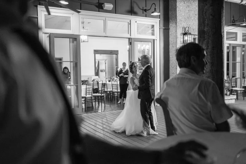 Black and white shot of Emma in her white dress standing with Kyle indoors, framed through a doorway with guests visible.