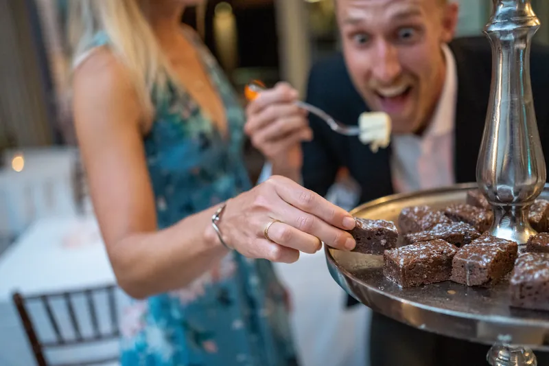 Two guests reach for chocolate desserts from a tiered stand, laughing and smiling during the reception.