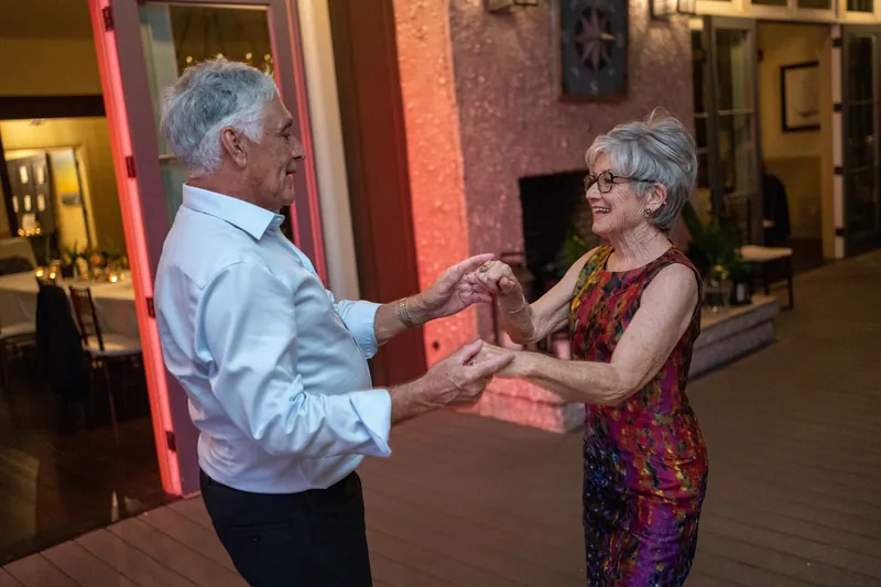 Two older guests dance together, holding hands with genuine smiles on an indoor dance floor.