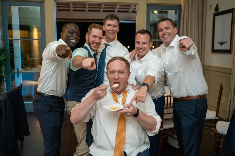 A group of five men in white shirts playfully pose together, with one man in the center biting an orange tie.
