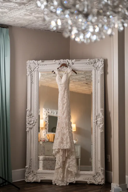 A lace wedding gown hangs in an ornate white frame against a taupe wall at The White Room, St. Augustine.