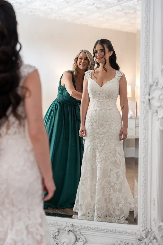 A person in a white lace gown stands with a bridesmaid in a teal dress, both reflected in an ornate mirror.