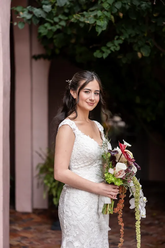 A person in a beaded white gown holds a cascading bouquet with pink and greenery against ivy-covered architecture.