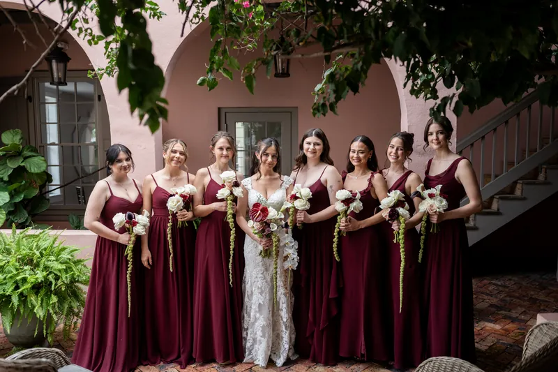 Eight people in burgundy bridesmaid dresses and one in a white gown stand together in a courtyard at The White Room.
