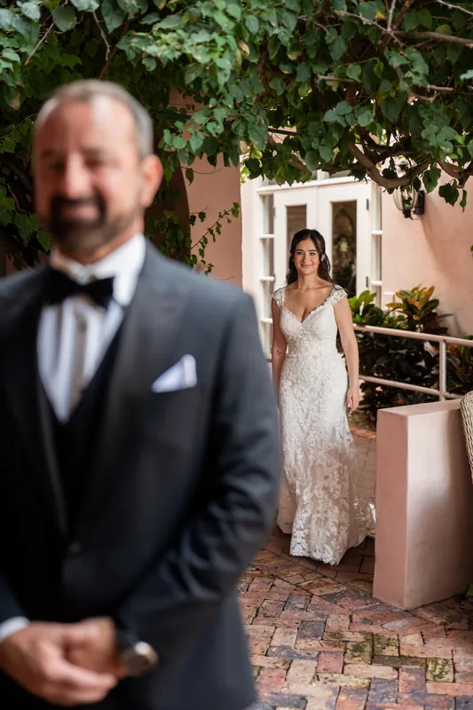 Two people in wedding attire stand in a brick courtyard; one in a dark suit is in sharp focus while the other in a white gown is in the background.