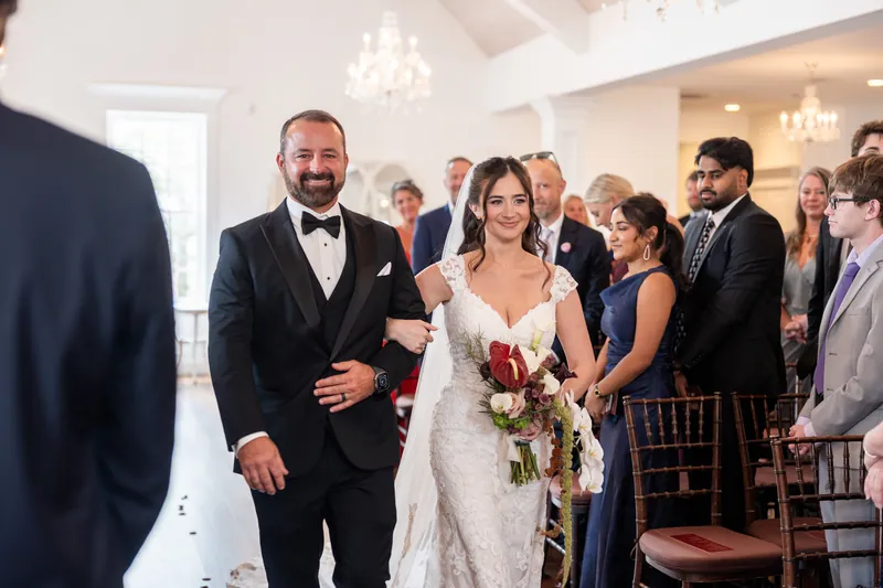 A couple walks down an aisle together during a ceremony, surrounded by seated guests in an elegant white hall.