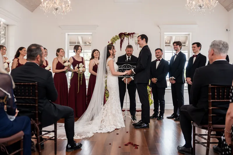 Emme and Andrew exchange vows at the altar under a floral arch as guests seated in burgundy and black look on at The White Room.