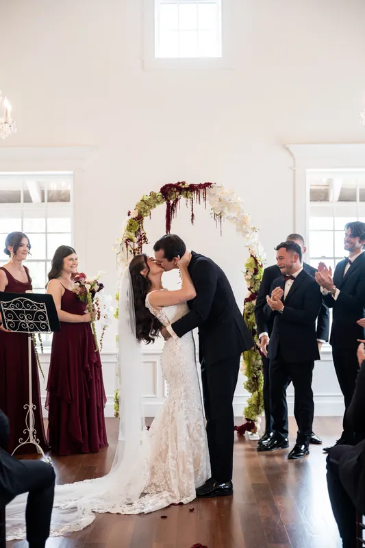 Emme and Andrew kiss under a draped floral arch while wedding guests applaud around them in the bright ceremony space.