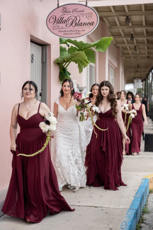 Bridesmaids in burgundy walk alongside Emme in her lace gown down a brick pathway outside the historic venue.