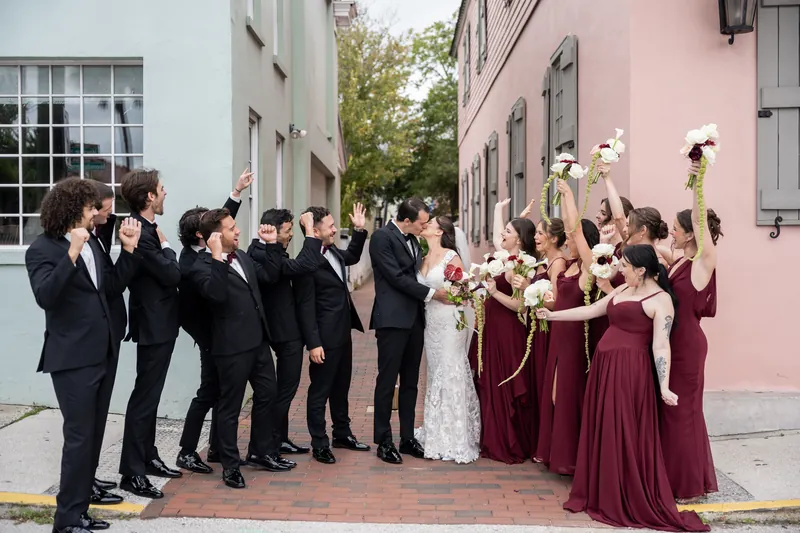 Emme and Andrew pose with their wedding party on a brick courtyard bordered by colonial architecture, guests cheering.