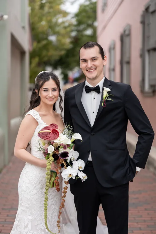 Emme and Andrew smile together on a brick alley, he in a tuxedo and she in a white lace gown holding burgundy and white flowers.