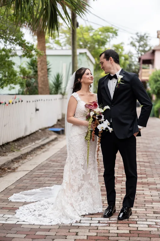 Two people stand on a palm-lined brick courtyard; the person in white holds a cascading bouquet while looking at their partner in a tuxedo.