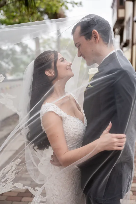 Beneath a flowing veil, two people gaze at each other on The White Room's brick terrace, their hands clasped as tropical greenery frames the scene.