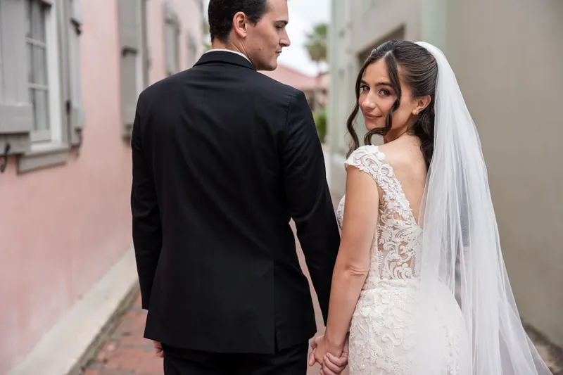 A person in a white lace gown and veil holds hands with their partner in a black tuxedo as they walk down a pink-walled alley.