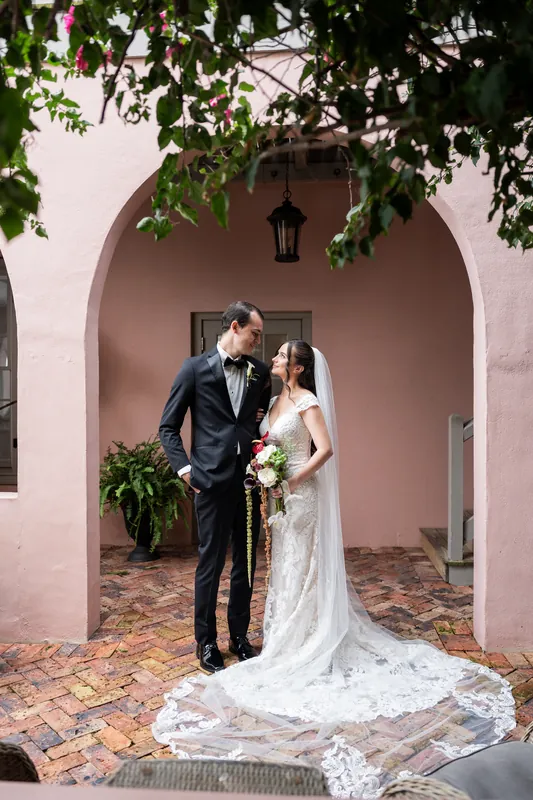 Under a pink arched entryway draped with green foliage, two people in wedding attire stand close together, the one in white holding a bridal bouquet.