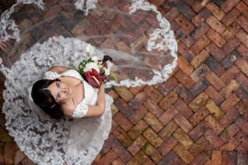Overhead view of a person in a white gown and veil on herringbone brick, holding a red and white bouquet and gazing down.