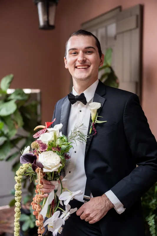 Andrew smiles in a black tuxedo with a white bow tie, holding a bridal bouquet with white roses and burgundy accents at The White Room.
