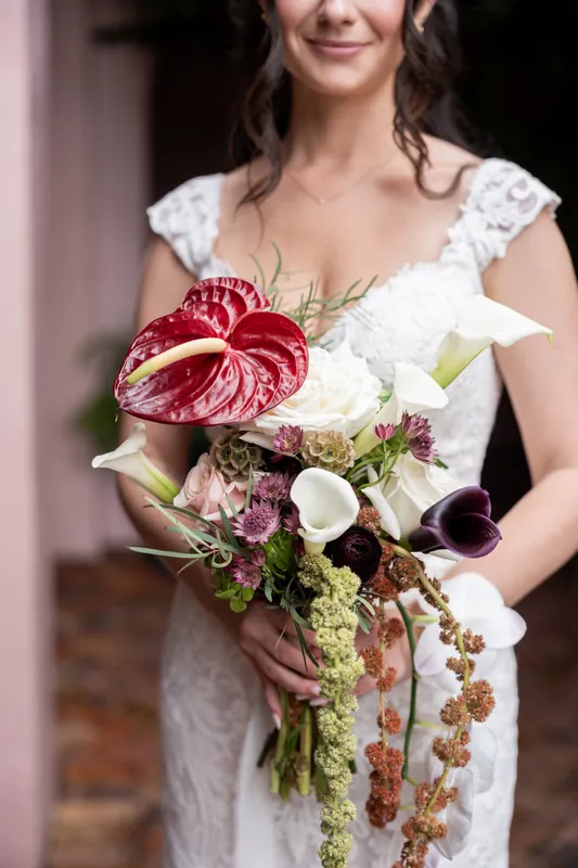 Someone in a white lace wedding gown holds a dramatic bouquet featuring deep red anthuriums, white calla lilies, and cascading foliage.