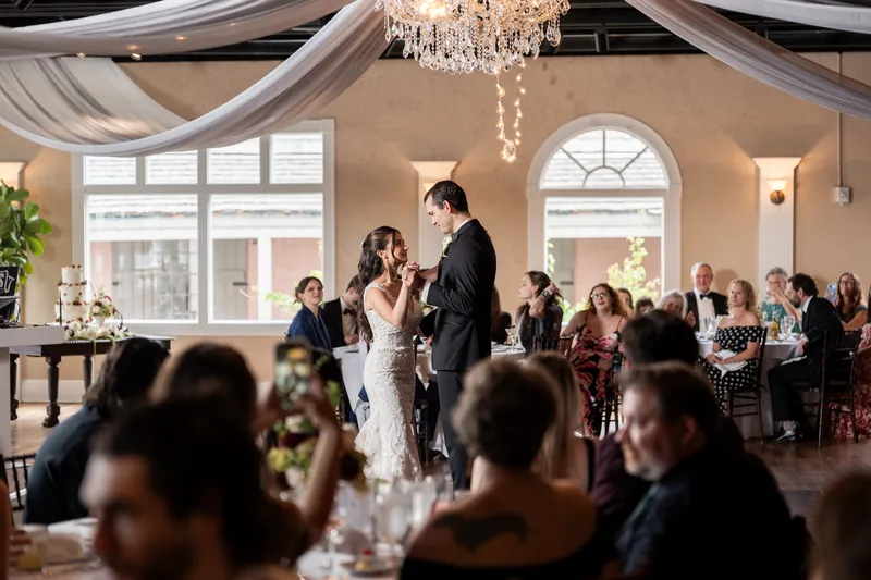 The couple stands together at the center of the reception hall under draped ivory fabric and a crystal chandelier, surrounded by seated guests.