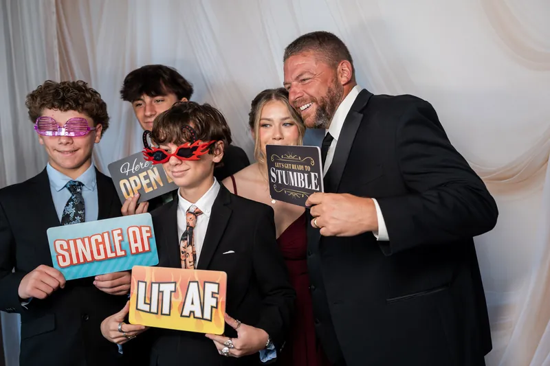 An adult and three children hold colorful photo booth props—including signs reading "Single AF" and "It AF"—at the wedding celebration.