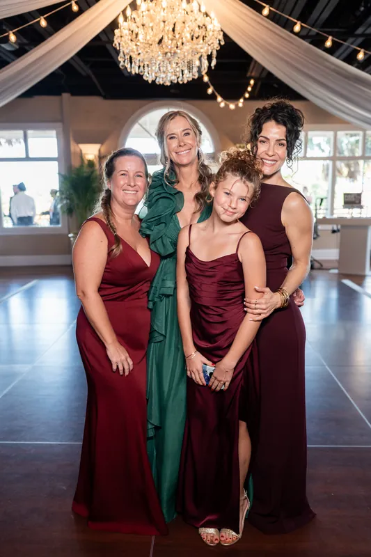 Three women and a young child pose together in burgundy and jewel-toned evening gowns beneath the chandelier in The White Room's ballroom.