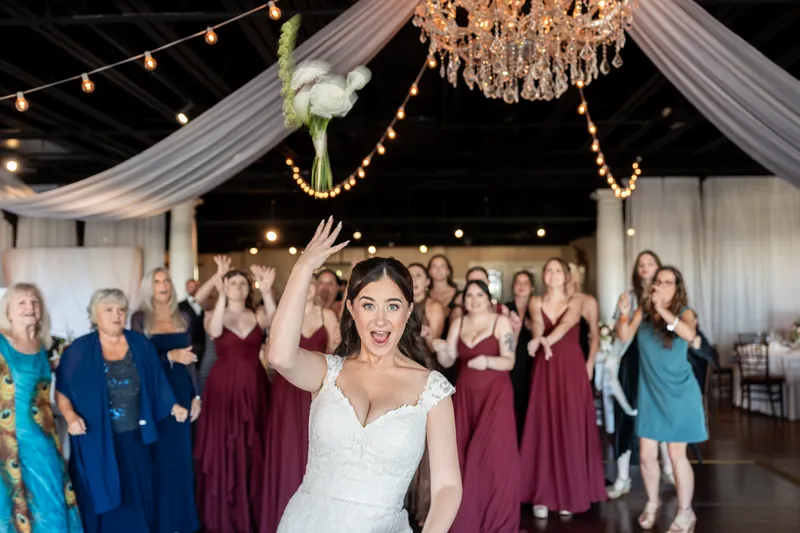 A person in a white gown tosses her bouquet toward guests in burgundy and blue dresses beneath a crystal chandelier at The White Room.