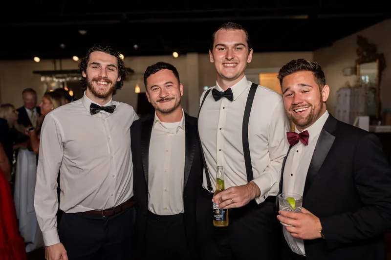 Four men in formal attire stand together holding drinks at The White Room's reception, smiling at the camera.
