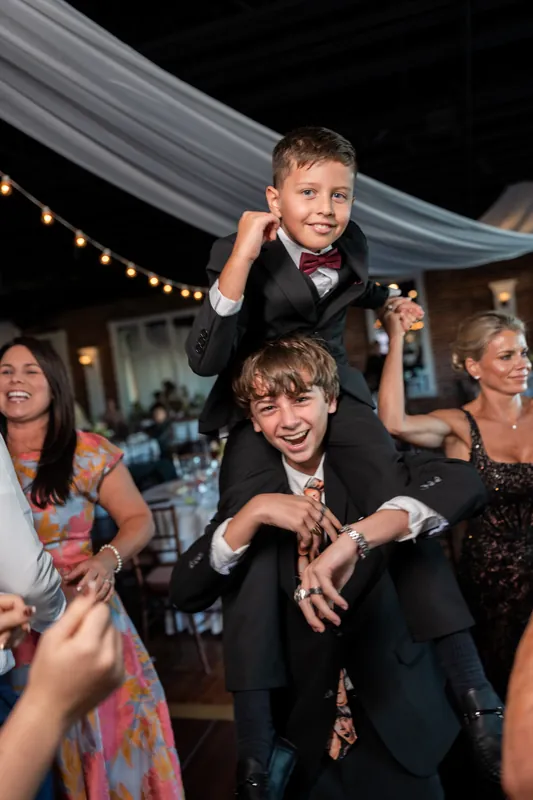 Two young boys in suits dance with adults during the reception at The White Room, one being lifted onto shoulders amid the crowd.