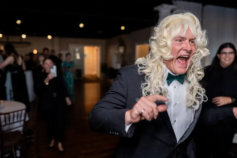 A guest in a navy suit and blonde curled wig points playfully at the camera while dancing at The White Room's reception.
