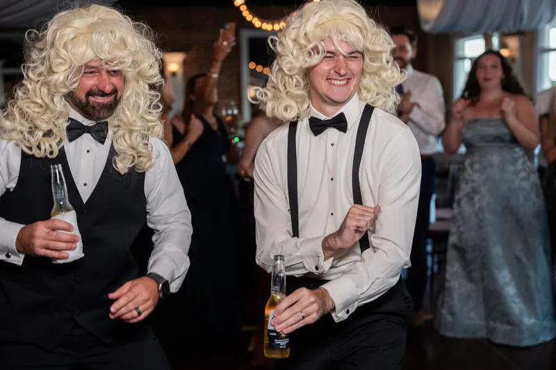 Two guests wearing matching blonde wigs and bow ties laugh together while holding drinks on the dance floor at The White Room.