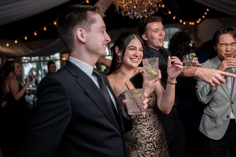 Two people in dark formal wear clink glasses mid-celebration while guests nearby raise their drinks under warm overhead lights.