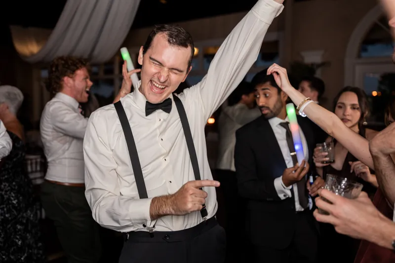 Someone in a white dress shirt celebrates with arms raised, surrounded by guests holding drinks and glow sticks at The White Room.