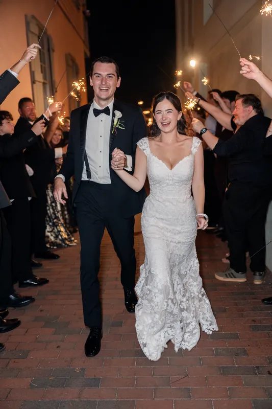 Emme and Andrew walk side by side through an outdoor brick corridor lined with guests waving sparklers, exiting The White Room.