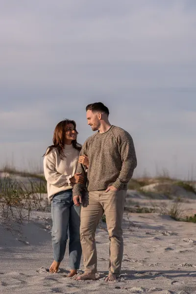 Eric and Anastasia share an intimate moment on a sandy beach during golden hour, gazing lovingly at each other while walking barefoot.