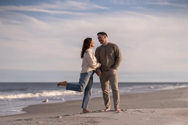 Eric and Anastasia share an intimate moment on the beach, with Anastasia playfully lifting her leg as they gaze into each other's eyes against the ocean backdrop.