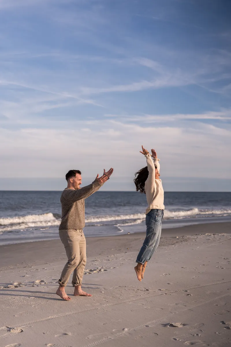 Eric and Anastasia share a joyful high-five moment on the beach, with Anastasia jumping up to meet his hands against the backdrop of ocean waves and cloudy sky.