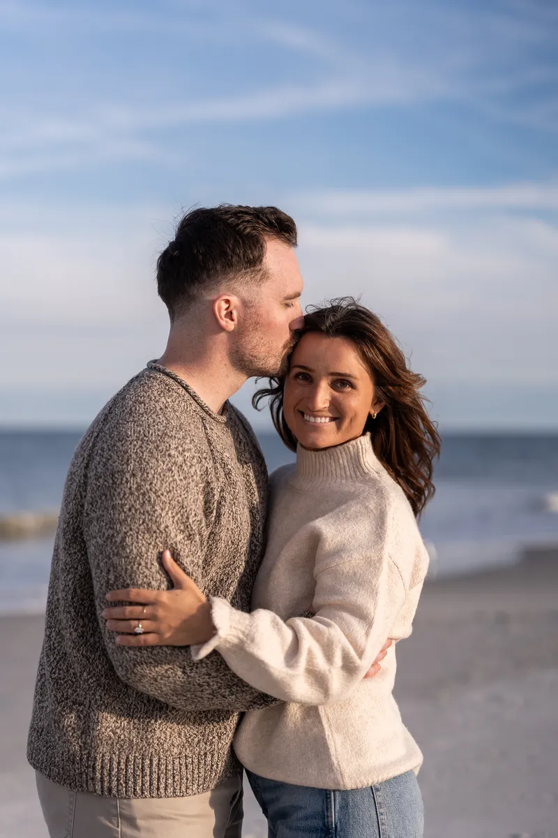 Eric and Anastasia share an intimate embrace on the beach, with him gently kissing her forehead as she smiles warmly at the camera.