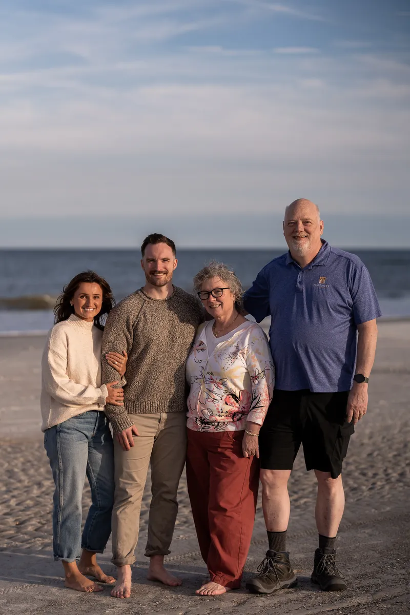A happy family of four poses together on a beach during golden hour, with the ocean and cloudy sky creating a beautiful backdrop.