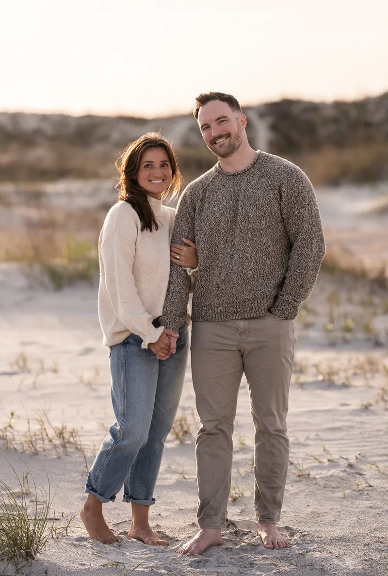 Eric and Anastasia share a joyful moment on the beach during golden hour, standing barefoot in the sand while holding hands and smiling at the camera.