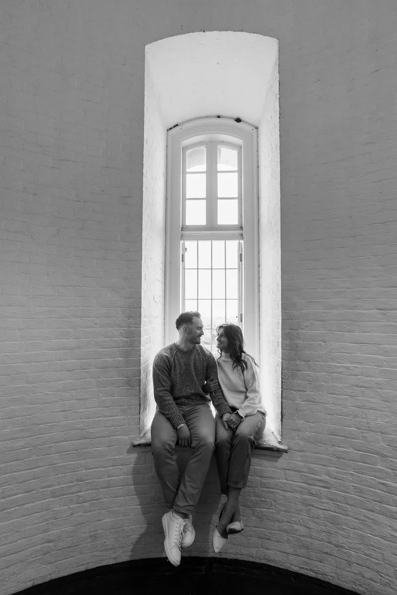 Eric and Anastasia sit together in a tall arched window alcove, sharing an intimate moment as they gaze at each other with warm smiles.