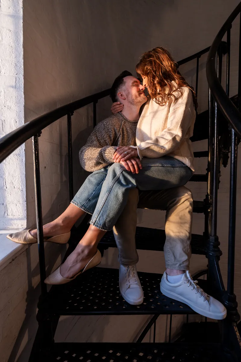 Eric and Anastasia share an intimate moment on a staircase, with warm golden light creating a romantic atmosphere as they gaze into each other's eyes.