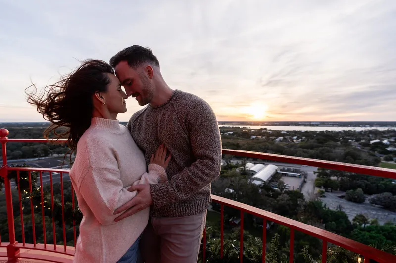 Eric and Anastasia share an intimate moment on a scenic overlook, embracing as the sun sets behind them over a beautiful landscape.