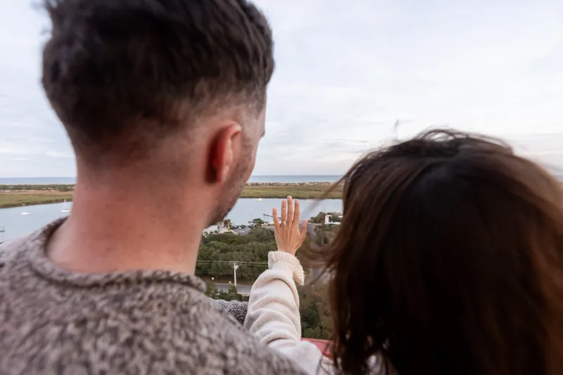 Eric and Anastasia share an intimate moment overlooking a scenic waterfront landscape, with Anastasia pointing toward the horizon as they take in the view together.