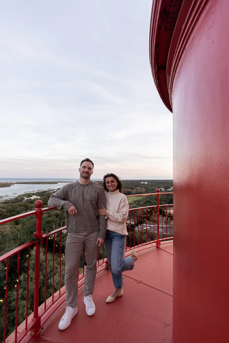 Eric and Anastasia pose together on a red lighthouse observation deck, enjoying golden hour views over the coastal landscape.