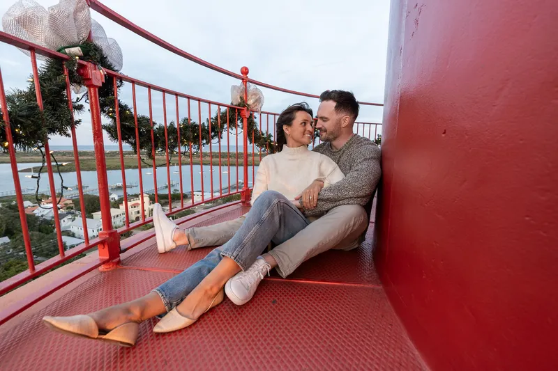 Eric and Anastasia share an intimate moment sitting together on a red lighthouse platform, gazing lovingly at each other during their engagement session.